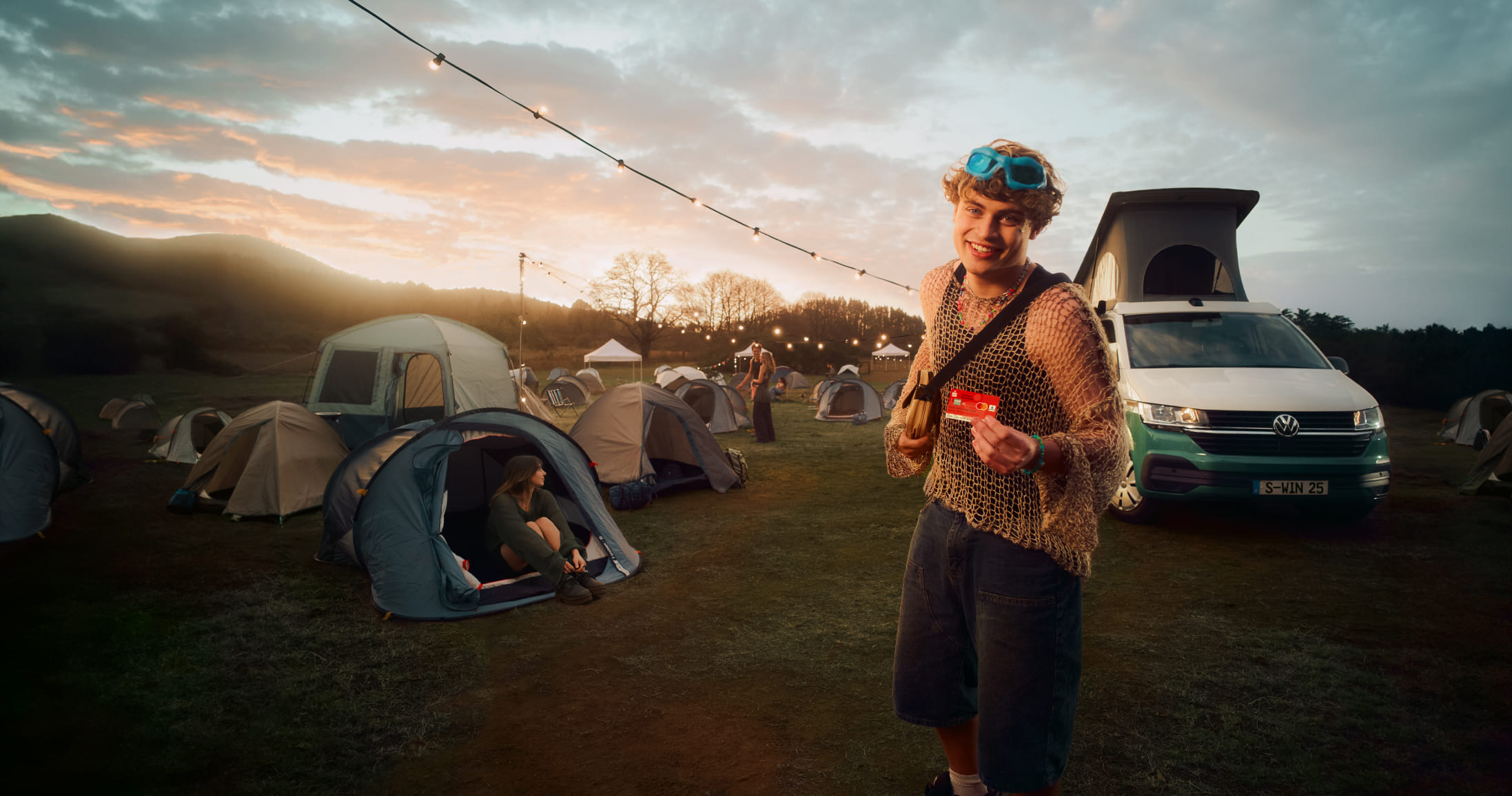 Ein junger Mann hält eine Sparkassenkarte in der Hand, während im Hintergrund ein VW Camper zu sehen ist, den man gewinnen kann.